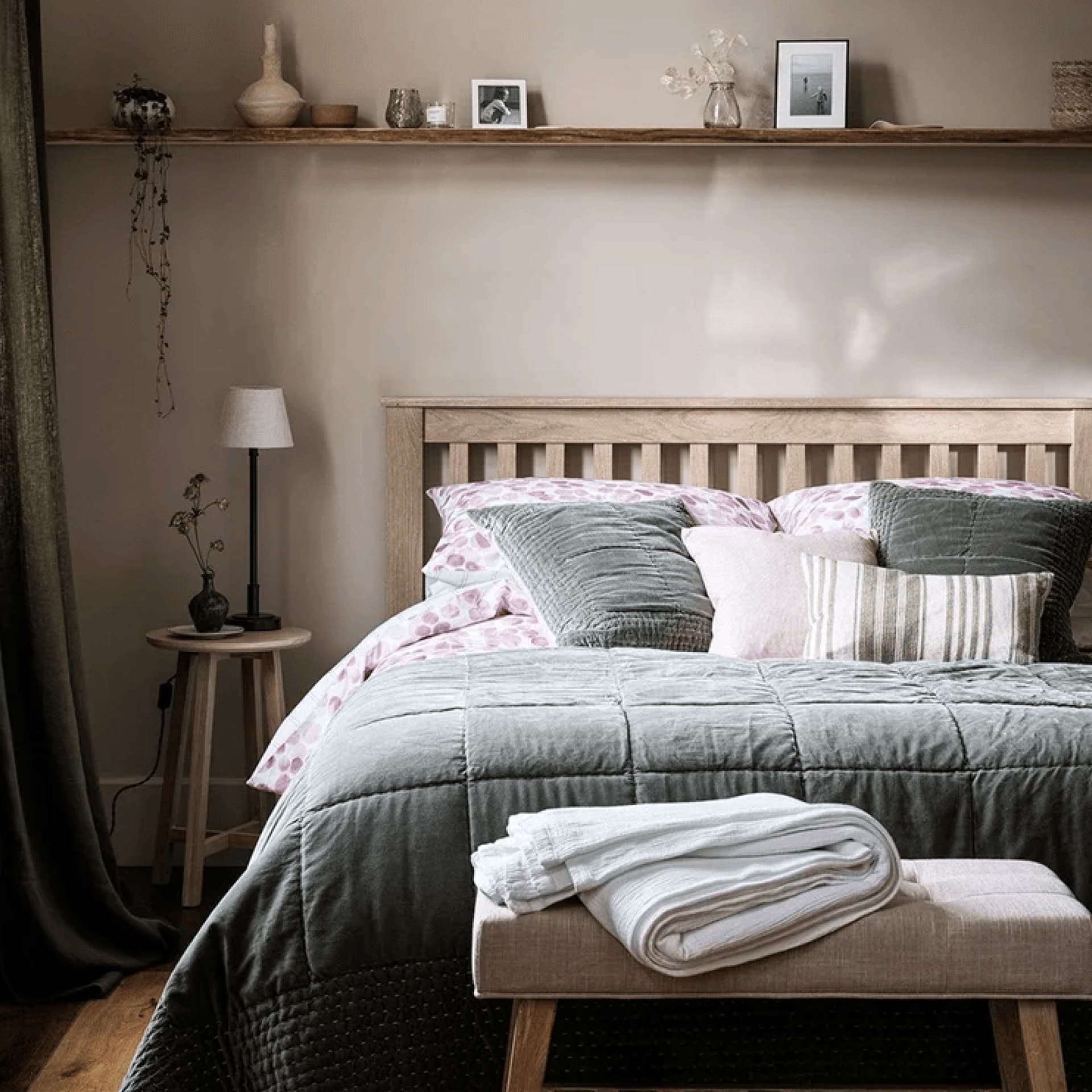 Cozy bedroom with a wooden headboard, pink and gray bedding, bedside table, and shelf with decor items. A bench with folded towels is in front.