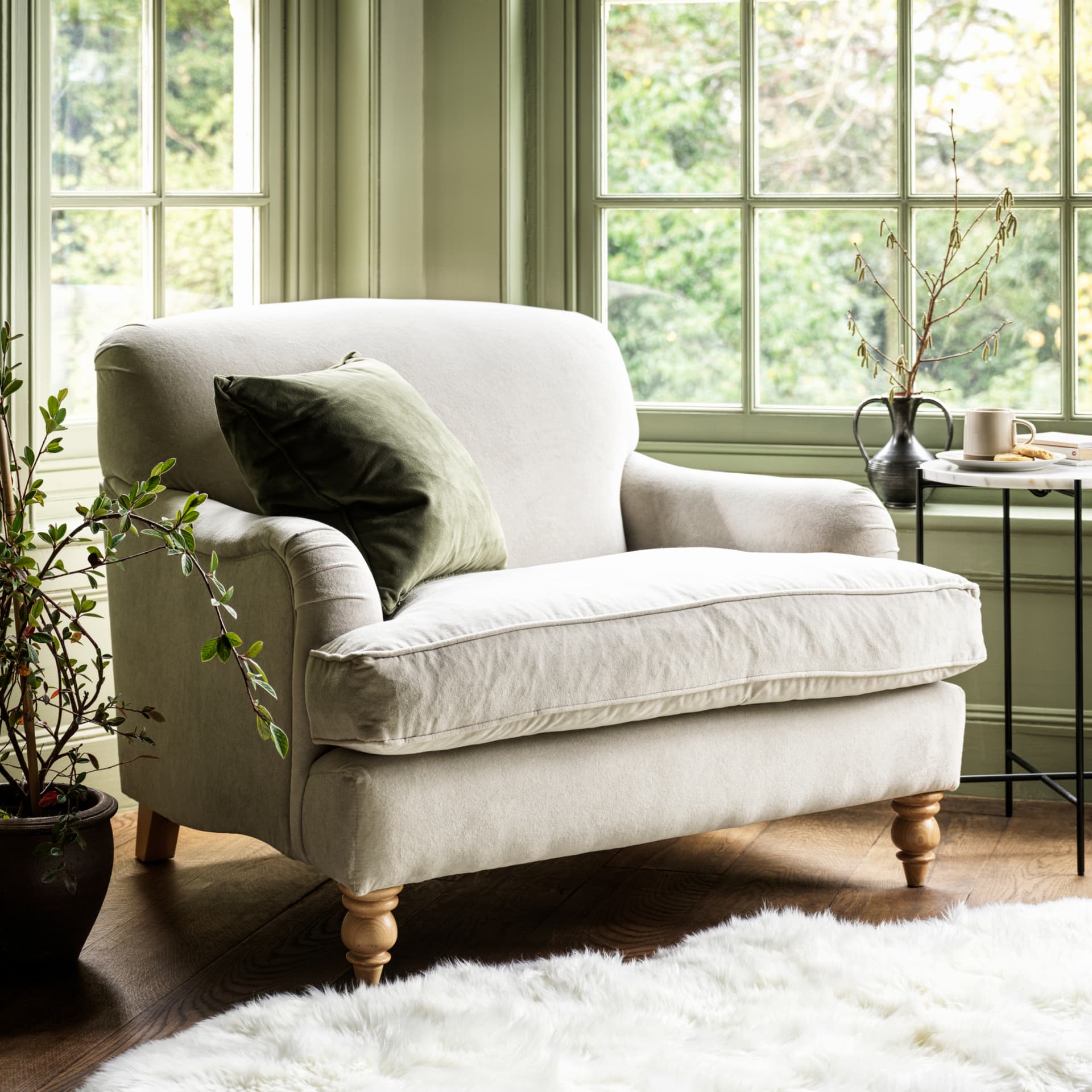 Cozy neutral armchair with a green throw pillow by large windows, surrounded by plants, a side table with a cup, and a fluffy white rug.