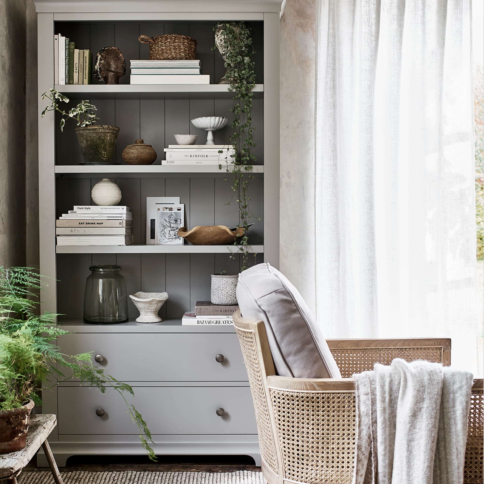 Cozy room with a grey bookshelf filled with books, plants, and decor. A wicker chair with a cushion and blanket sits by a bright window.