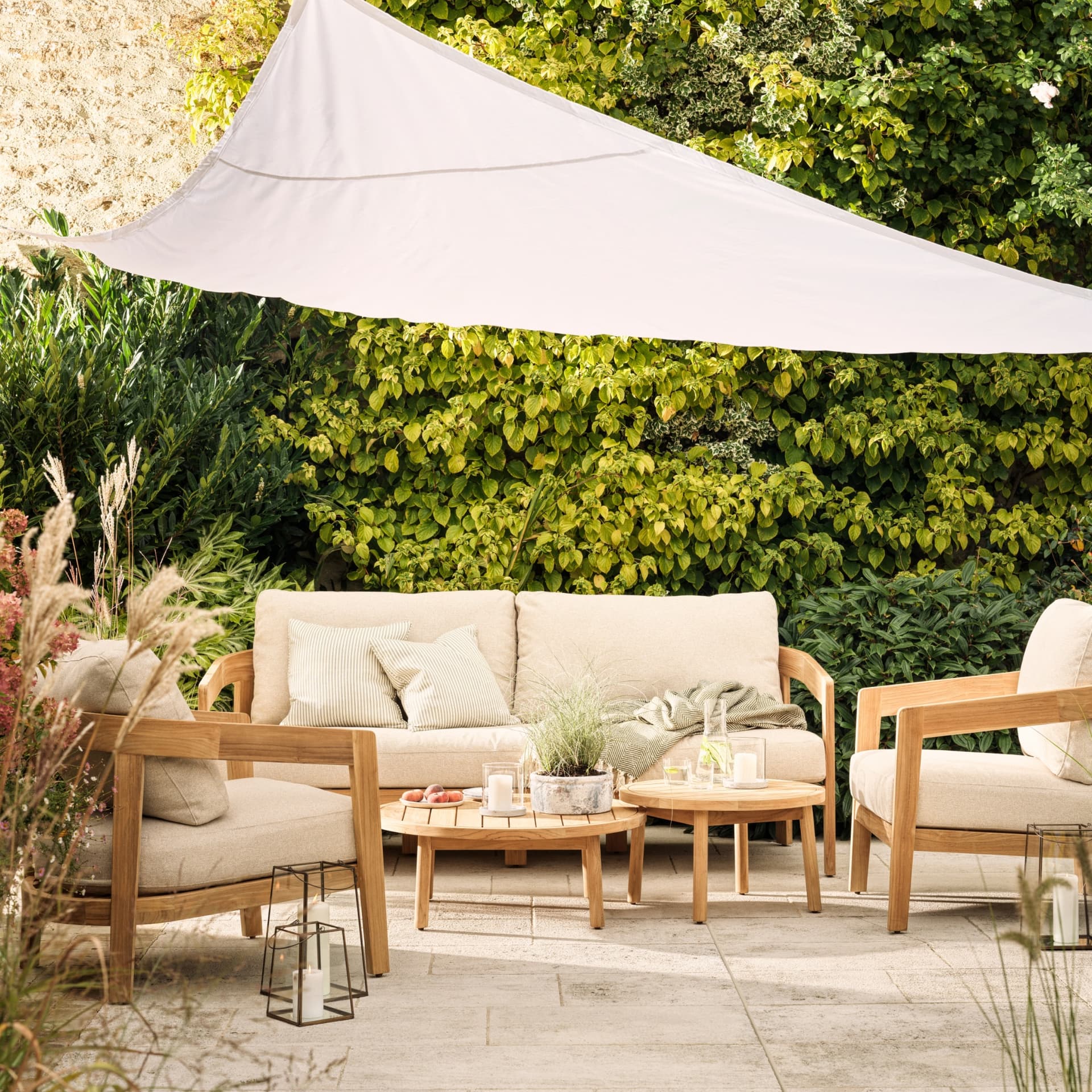 Outdoor patio with beige cushioned seating, wooden tables, candles, and greenery under a white sunshade.