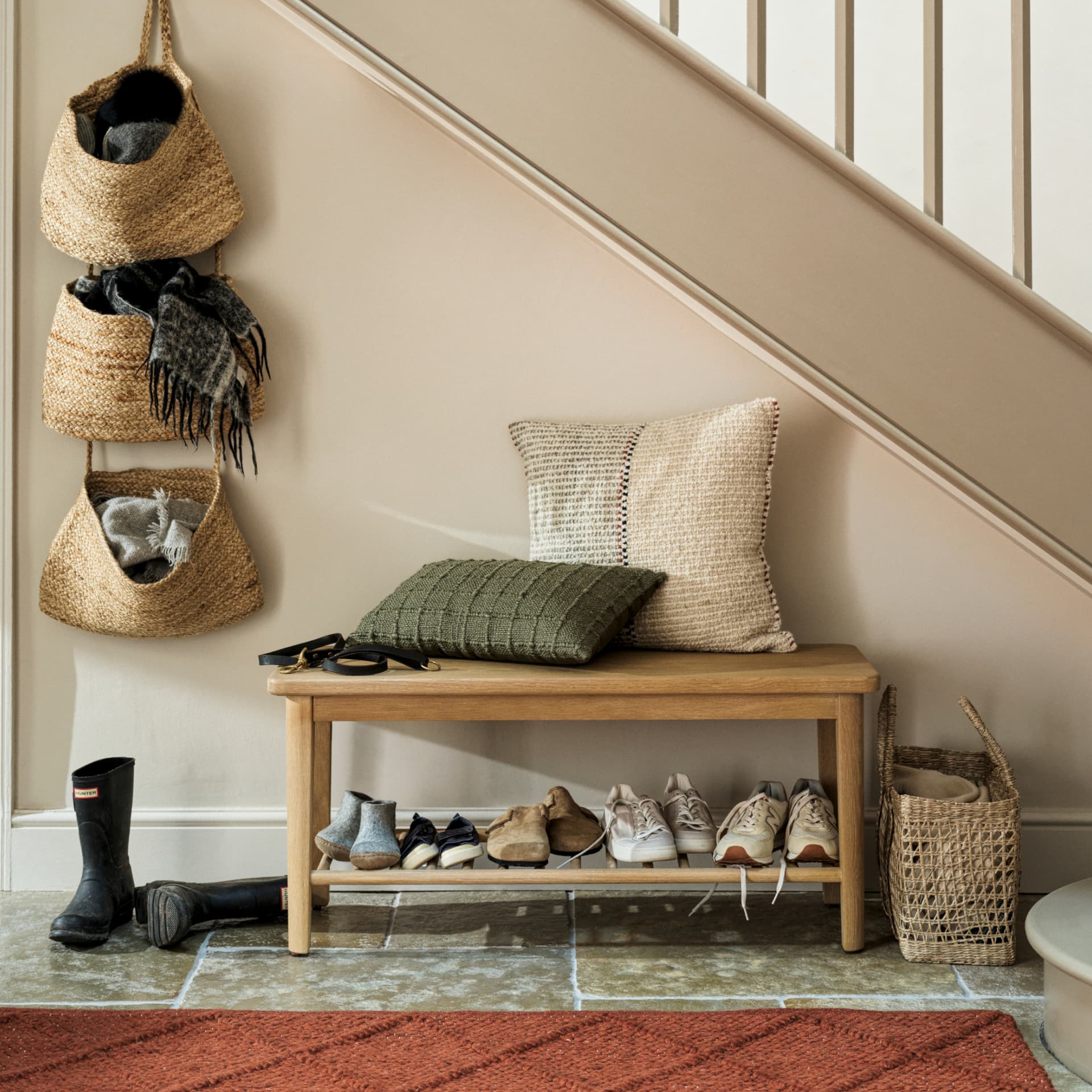 Cozy entryway with a wooden bench, cushions, shoes, and woven baskets holding scarves. Rubber boots are placed nearby on the stone floor.