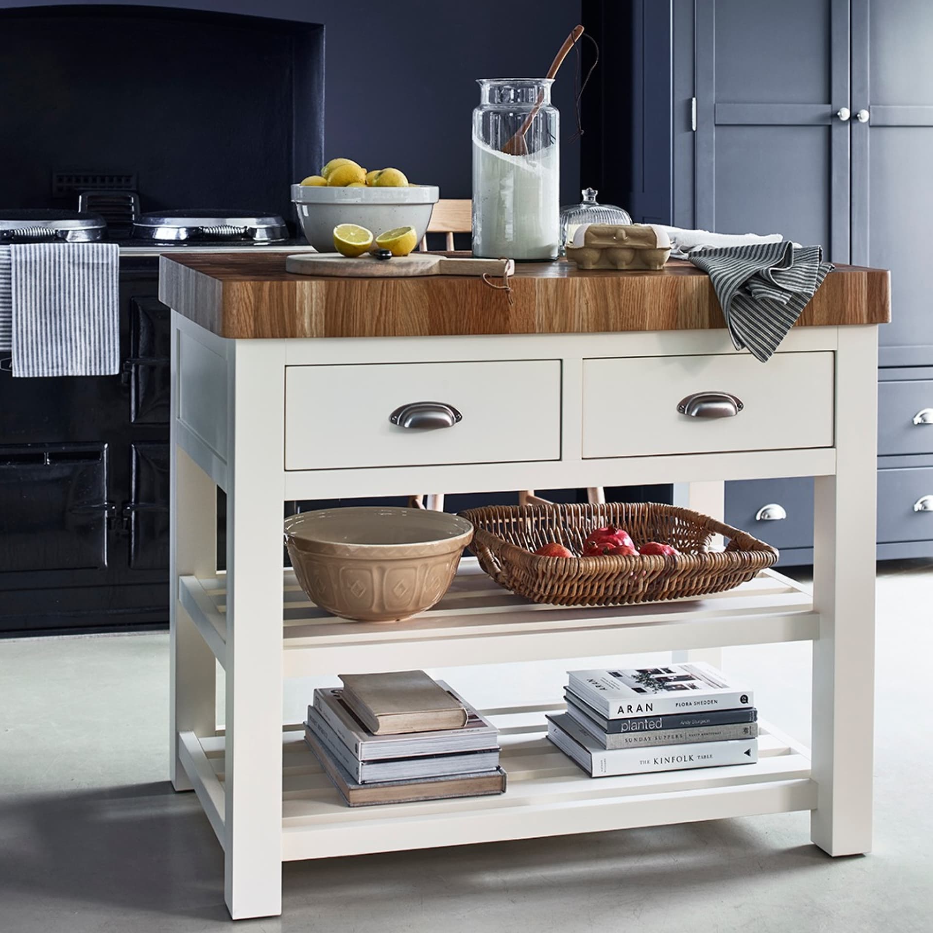 White kitchen island with a wooden top, lemons, a glass jar, eggs, striped cloth, onions in a basket, and books on the shelves below.