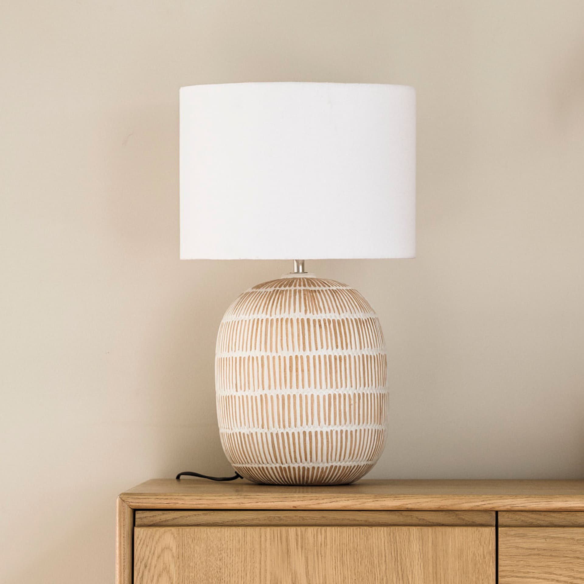 A ceramic table lamp with a white shade sits on a wooden surface. The base has a geometric pattern with beige and white stripes.