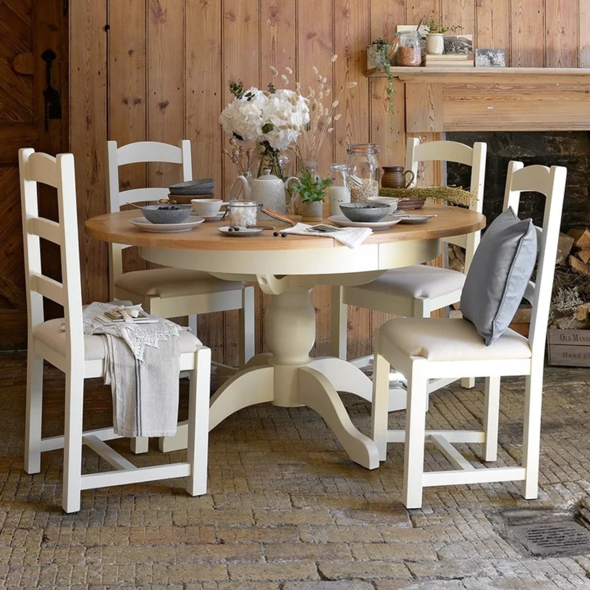 Rustic dining setup with a round wooden table, white chairs, floral centerpiece, and tableware, set in a cozy room with wood-paneled walls.
