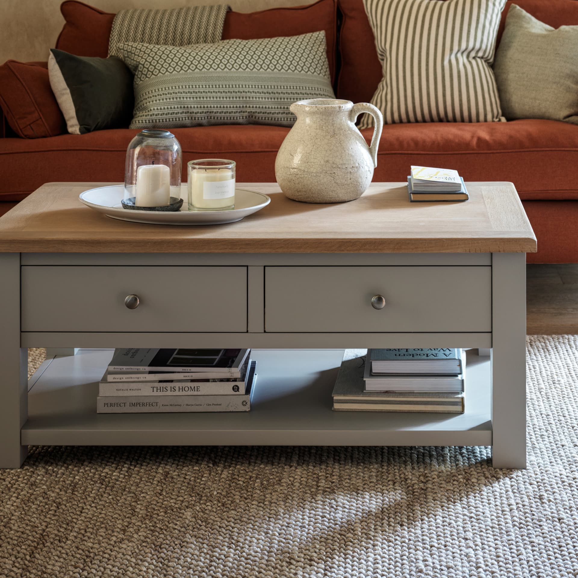 A living room with a wooden coffee table, candles, a ceramic jug, and books. A rust coloured sofa with patterned cushions is in the background.