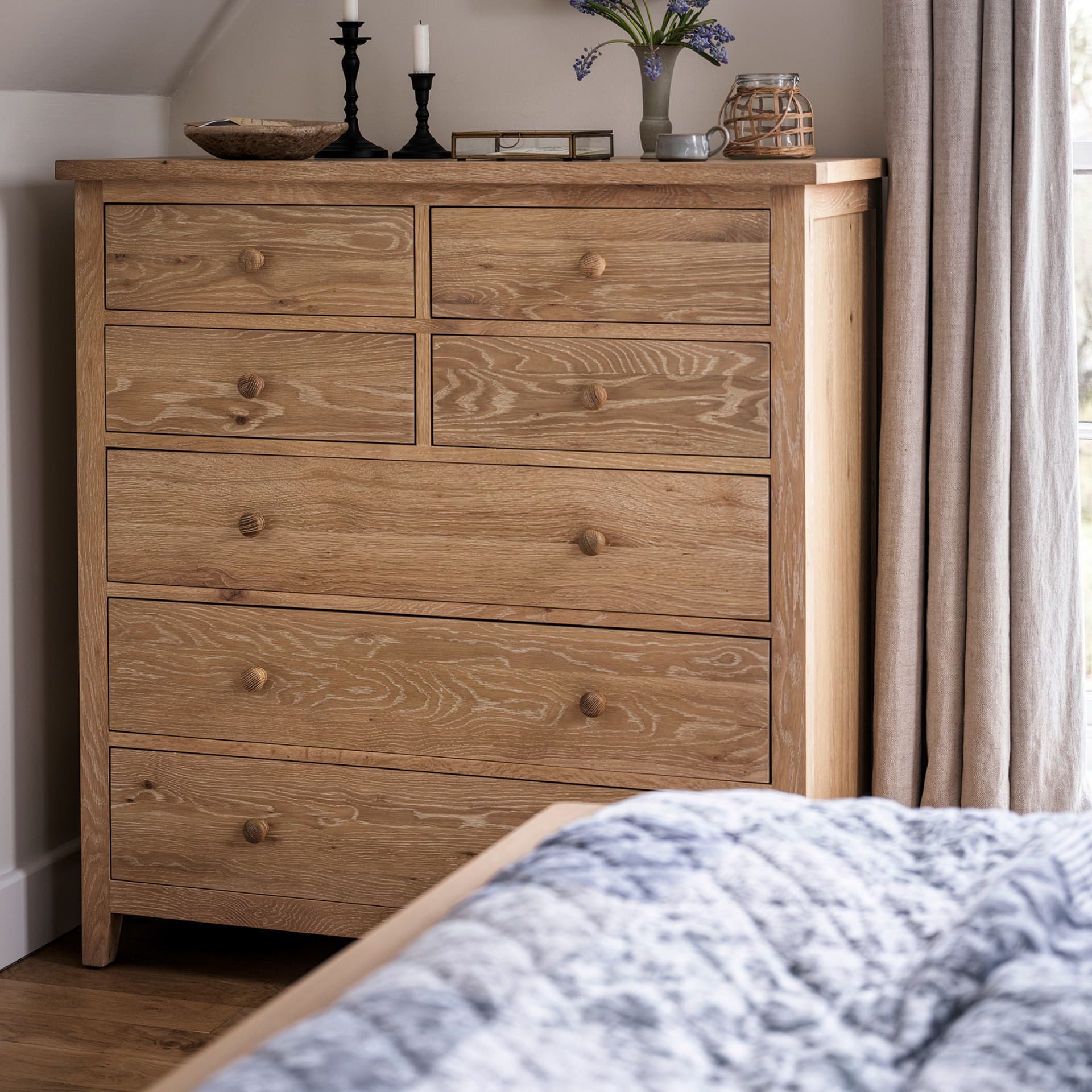 A light wooden dresser with seven drawers, topped with decorative items, stands beside a curtained window in a cozy bedroom setting.