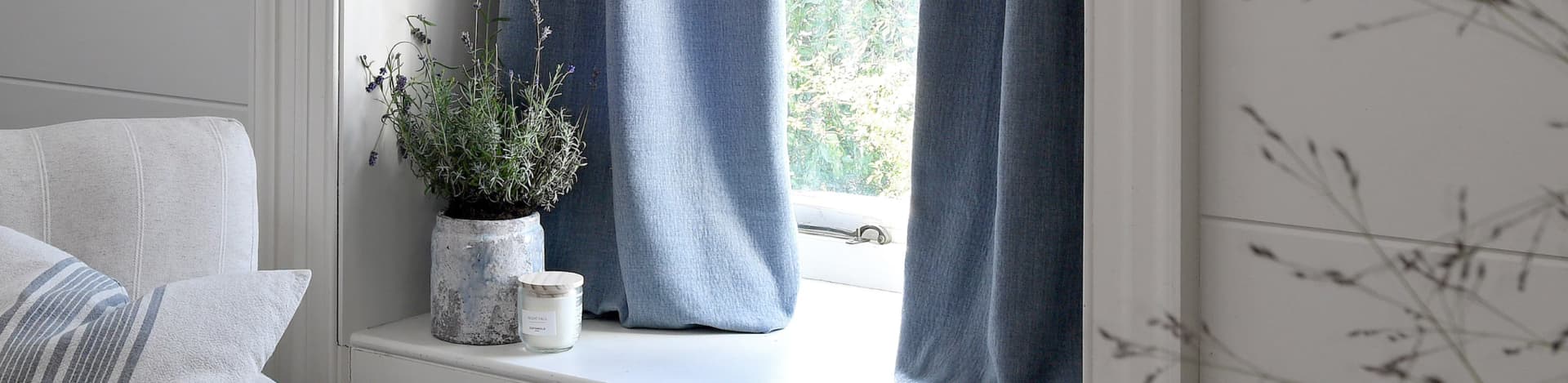 A cozy window nook with blue curtains, a potted lavender plant, and a lit candle, set beside a cushioned seat with a striped pillow.