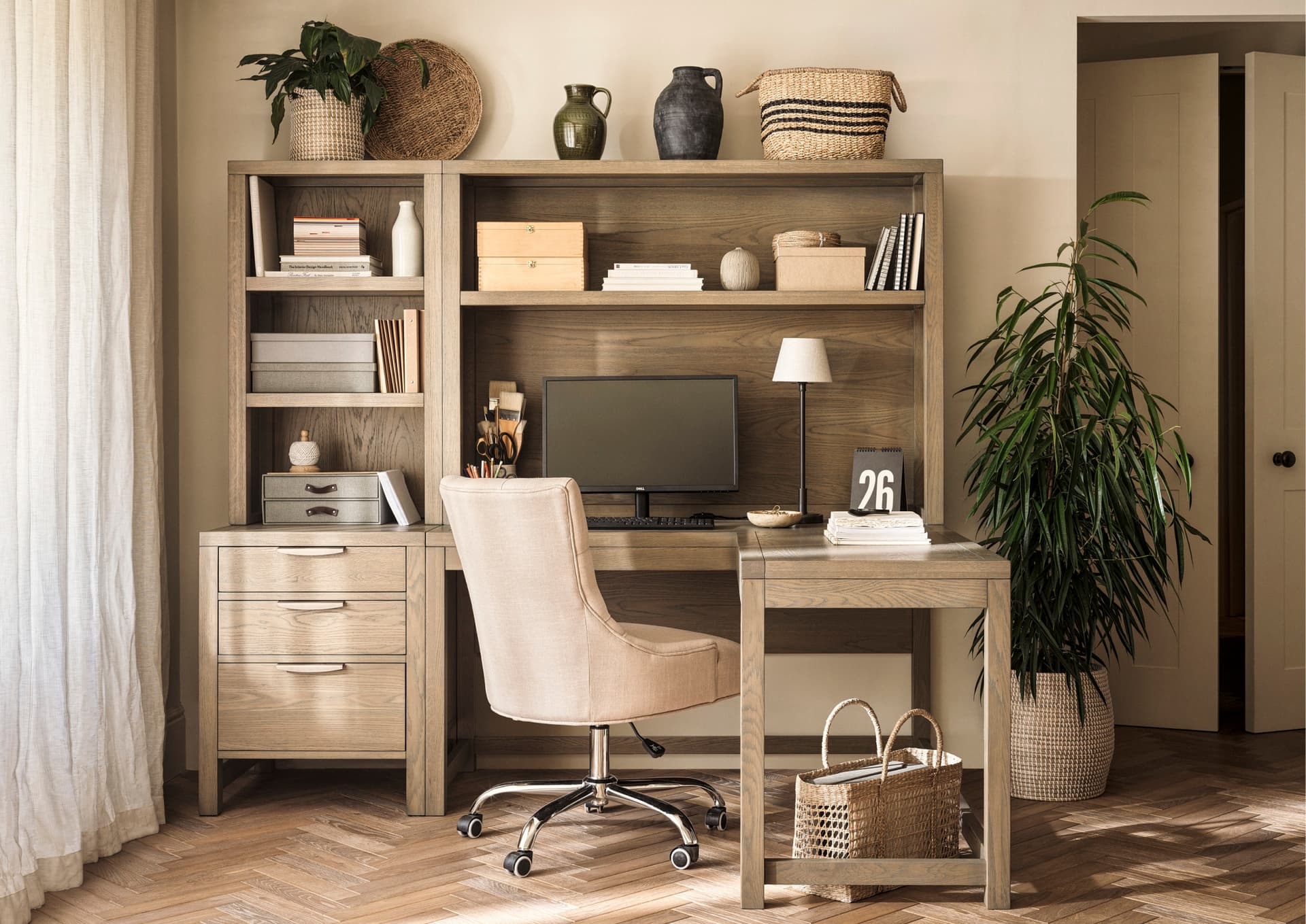 Home office with a wooden desk, beige chair, computer, potted plant, and shelves with decor, set in a room with herringbone-patterned flooring.