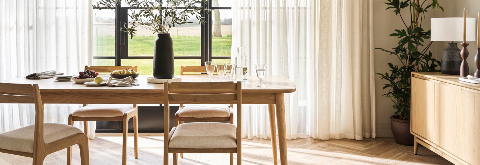 Modern dining room with a wooden table, chairs, and sideboard. Table set with plates, fruit, and wine glasses. Large windows and sheer curtains.