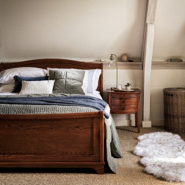 Cozy bedroom with a wooden bed, layered bedding, a side table with a lamp, wicker basket, and a sheepskin rug on a carpeted floor.