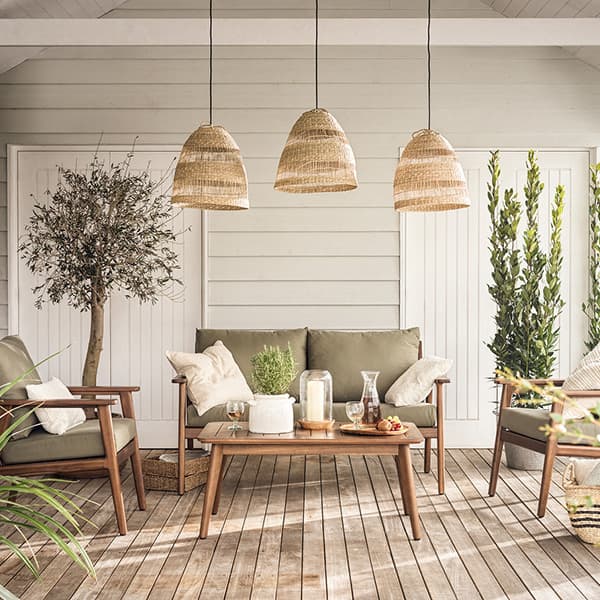 Cozy outdoor patio with wicker pendant lights, wooden sofas with green cushions, a wooden table, potted plants, and neutral decor under a wooden ceiling.