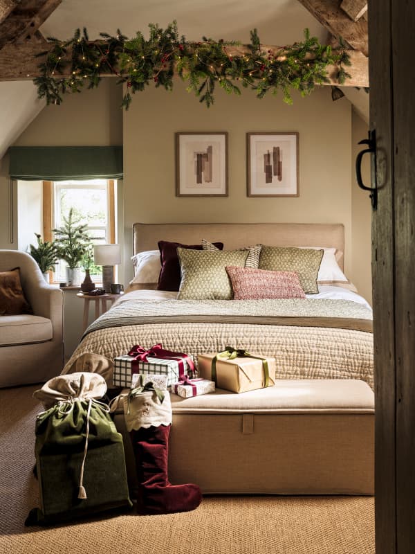 Cozy bedroom with a double bed, festive greenery above, presents and stockings on a bench, and a cushioned chair by the window.