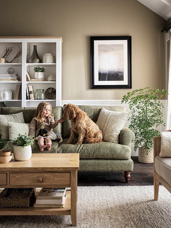 A cozy living room with a child and a dog on a green sofa, surrounded by plants and a wooden coffee table,