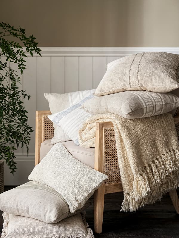 Beige and cream cushions stacked on a wooden chair with a cozy throw blanket, next to a potted plant against a paneled wall.