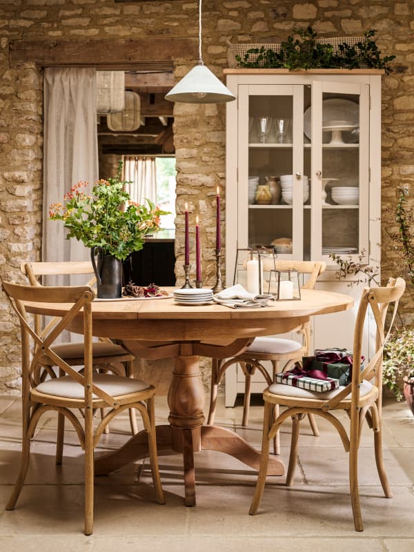 A rustic dining room with a wooden table set for four, surrounded by chairs, a vase of flowers, candles, and gifts, with a stone wall backdrop.