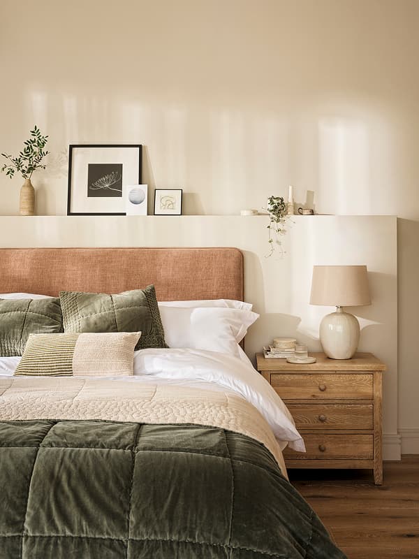 Cosy bedroom with an upholstered headboard, green velvet bedding, wooden nightstand with a lamp, framed art, and plants on a shelf above.