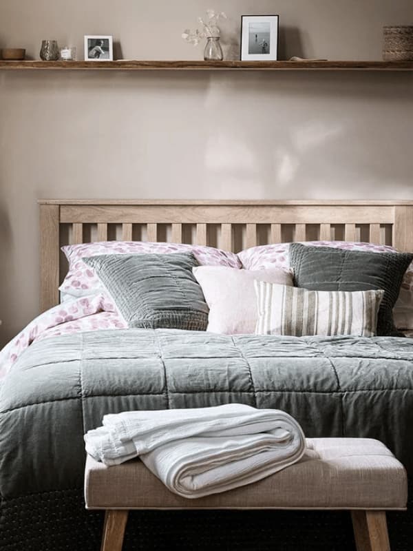 Cozy bedroom with a wooden headboard, gray and pink bedding, and pillows. A folded blanket sits on a bench, and a shelf displays photos.