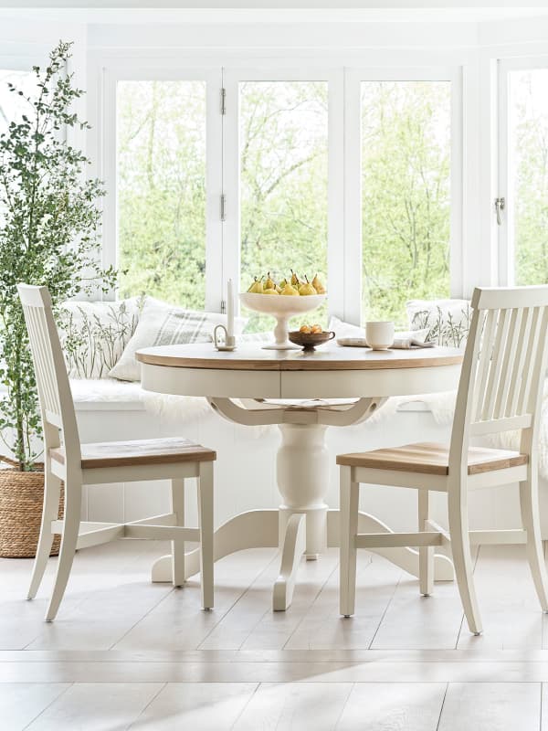 Bright dining area with a round wooden table, two chairs, and a window seat. Table features a fruit bowl and cups, set against large windows.