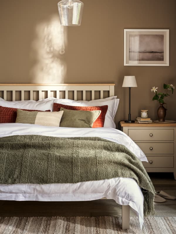 Bedroom with a wooden bed, green blanket, and assorted pillows. Warm lighting, wall art, and a nightstand with a lamp and vase.