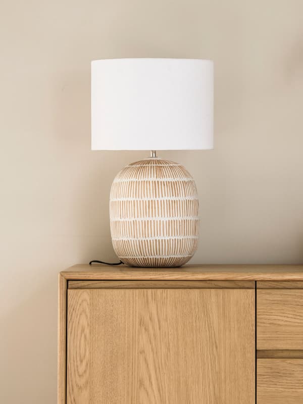 A ceramic table lamp with a white shade and a patterned beige base sits on a light wood cabinet against a neutral wall.