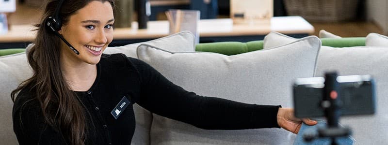 Smiling woman with a headset sitting on a couch, gesturing towards a smartphone on a tripod, engaging in a video call.