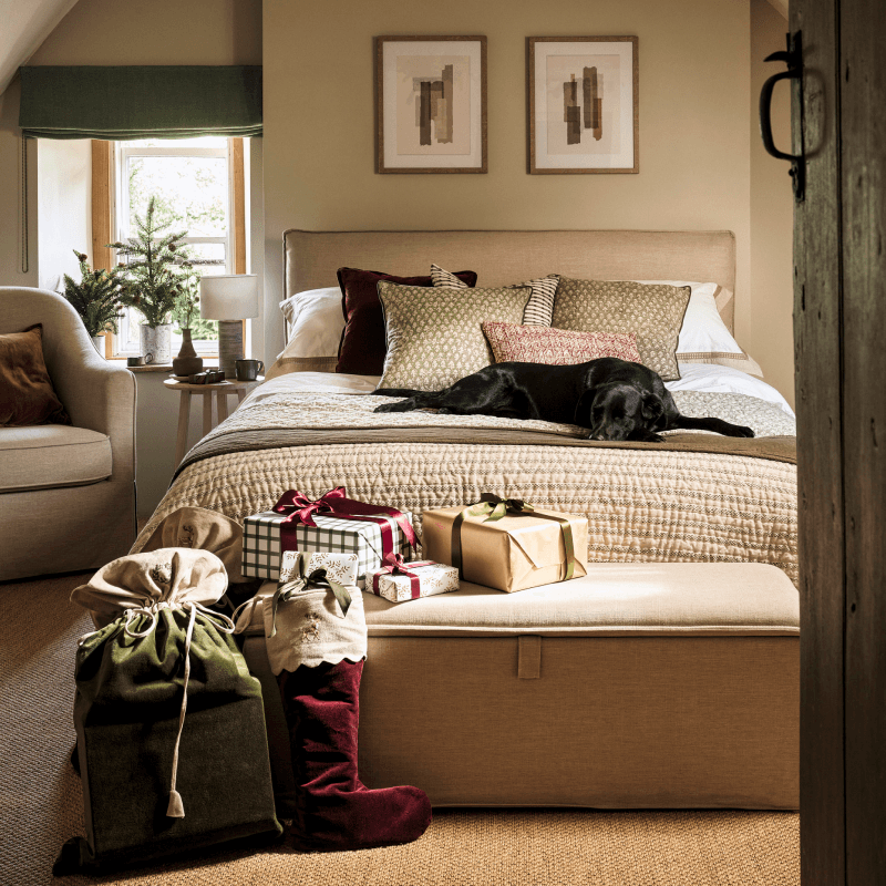 A cosy bedroom with a dog resting on a neatly made bed, surrounded by wrapped presents and festive decorations.
