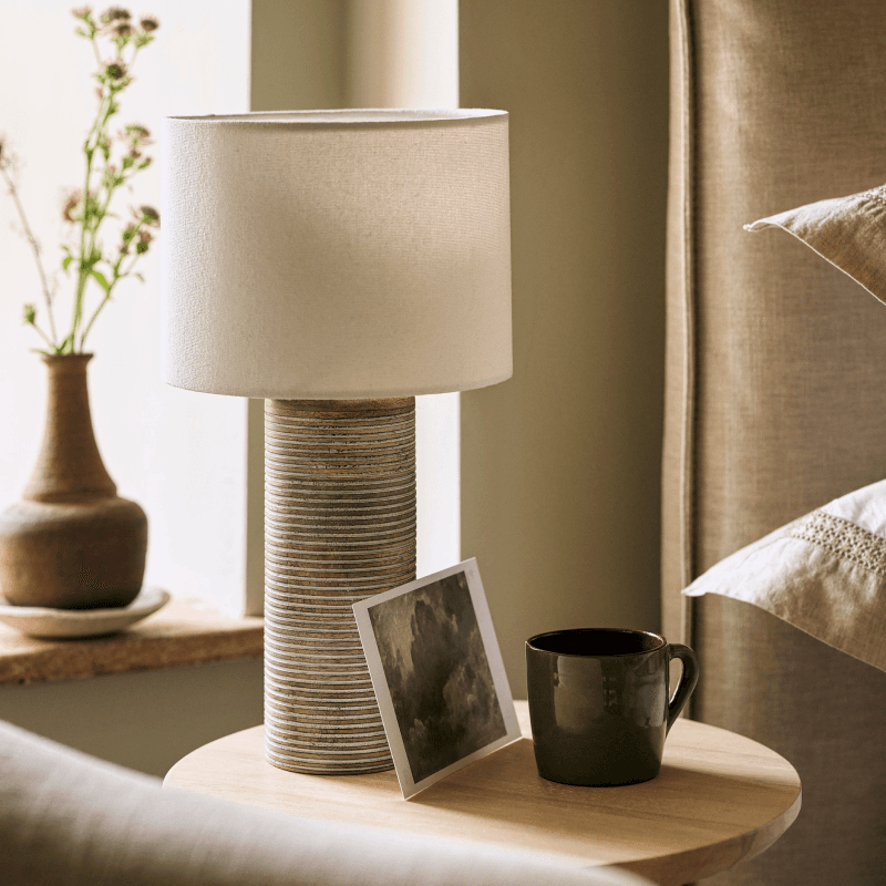 A cosy bedside scene with a textured lamp, a photo, and a dark mug on a wooden table, with a vase of dried flowers in the background.