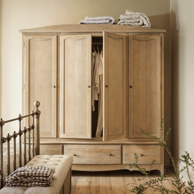 A wooden wardrobe with open doors revealing clothes, folded blankets on top, an upholstered bench in front, and a metal bed frame nearby.