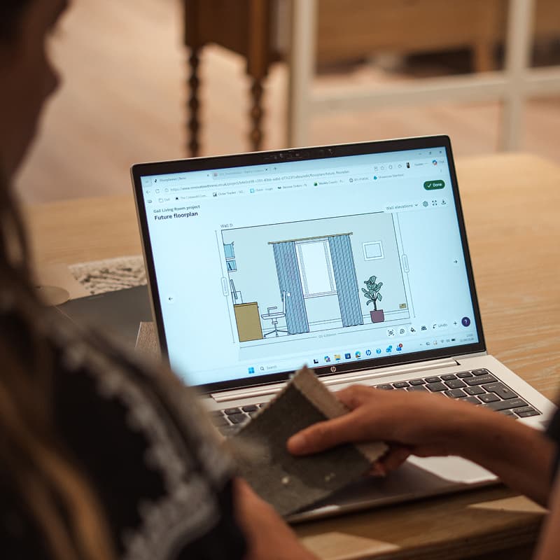 Person holding fabric samples, looking at a laptop displaying an interior design floor plan with curtains and furniture.