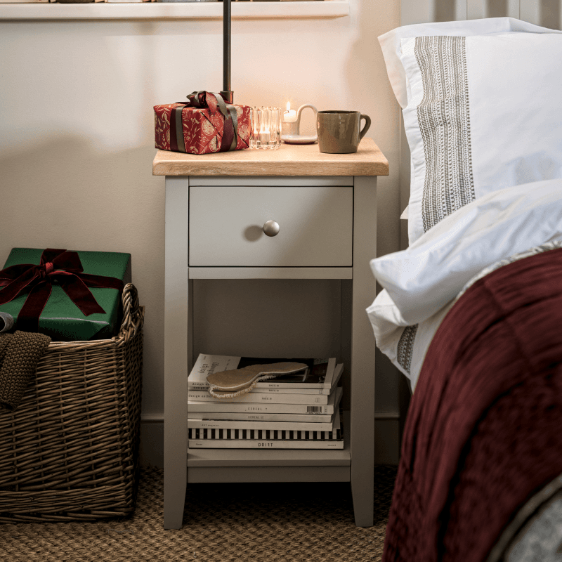 A cozy bedside table with a candle, mug, and wrapped gift on top, books below, next to a wicker basket with more gifts, beside a neatly made bed.