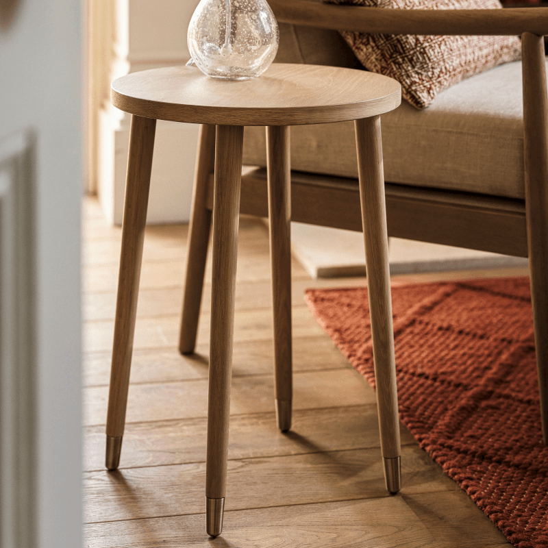 A wooden side table with tapered legs and a glass vase on top, next to a beige armchair with a patterned cushion, on a wooden floor.