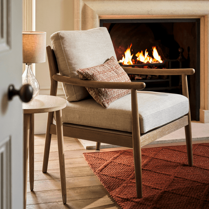 Cosy living room scene with a beige armchair, patterned red cushion, wooden side table with a lamp, and a lit fireplace in the background.