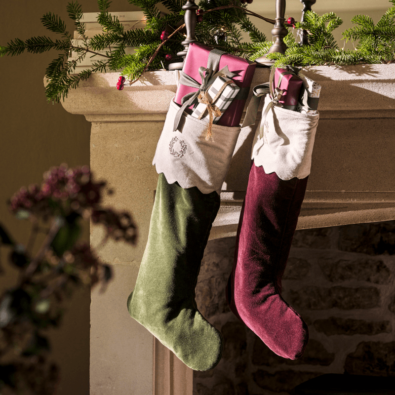 Two Christmas stockings, one green and one red, filled with wrapped gifts, hanging from a mantel decorated with greenery and berries.