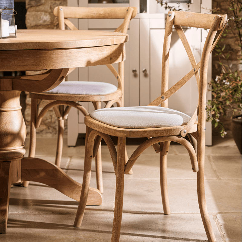 Dining room with round wooden table, two cross-back chairs with cushioned seats, stone floor, and a cabinet in the background.