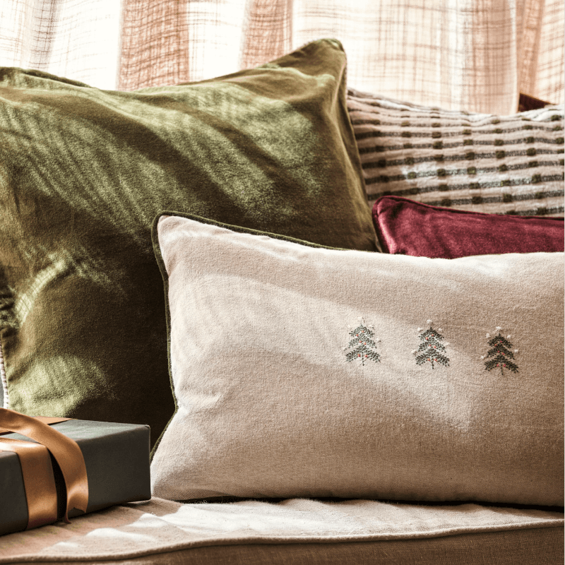 Cushions with varying textures and patterns, including a green velvet, maroon velvet, striped design, and a beige one with embroidered trees, arranged on a couch.