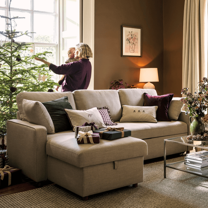 A cosy living room with a decorated Christmas tree by the window, a beige L-shaped sofa with pillows and wrapped gifts.