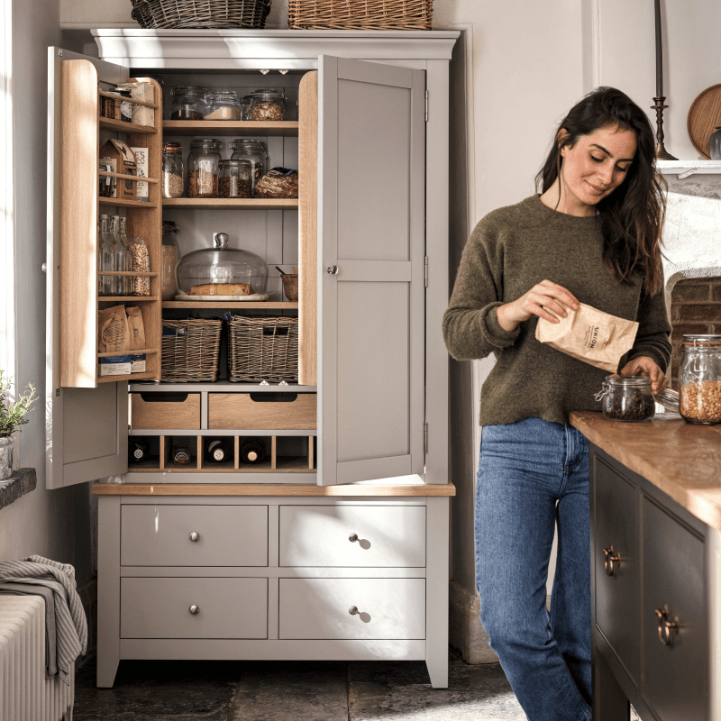 A woman in a cosy kitchen fills a jar with food from a pantry cupboard, which is open to display neatly organized jars and baskets.