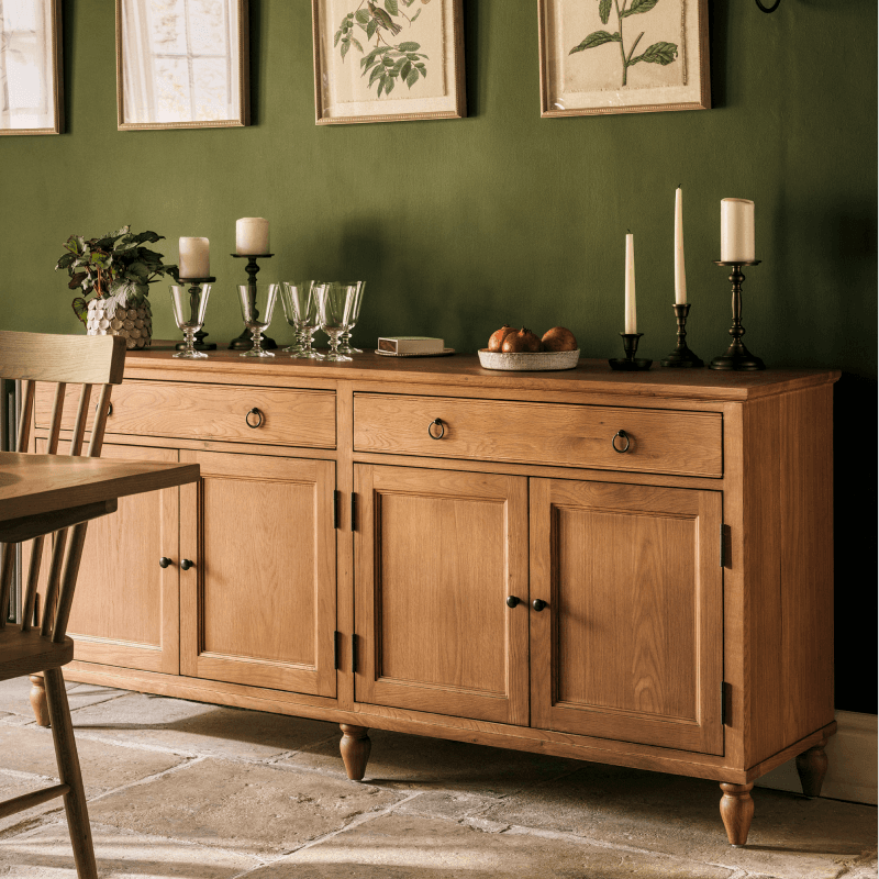 A wooden sideboard with candles, glassware, and fruit on top, set against a green wall with framed botanical prints above.