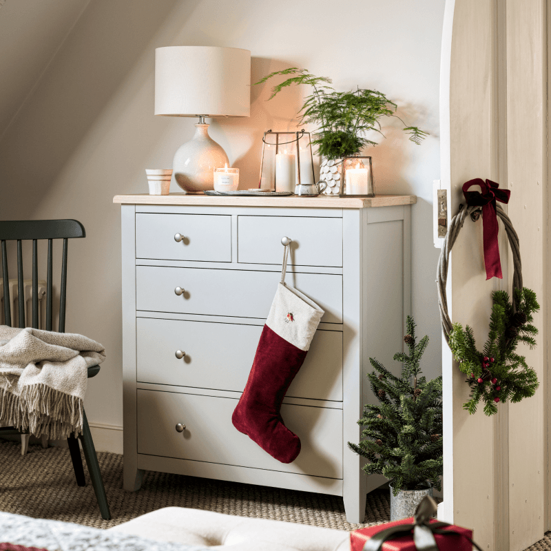 A cozy room featuring a light grey dresser with candles, a potted plant, and a lit lamp. A red stocking hangs on a drawer, and a wreath adorns the door.