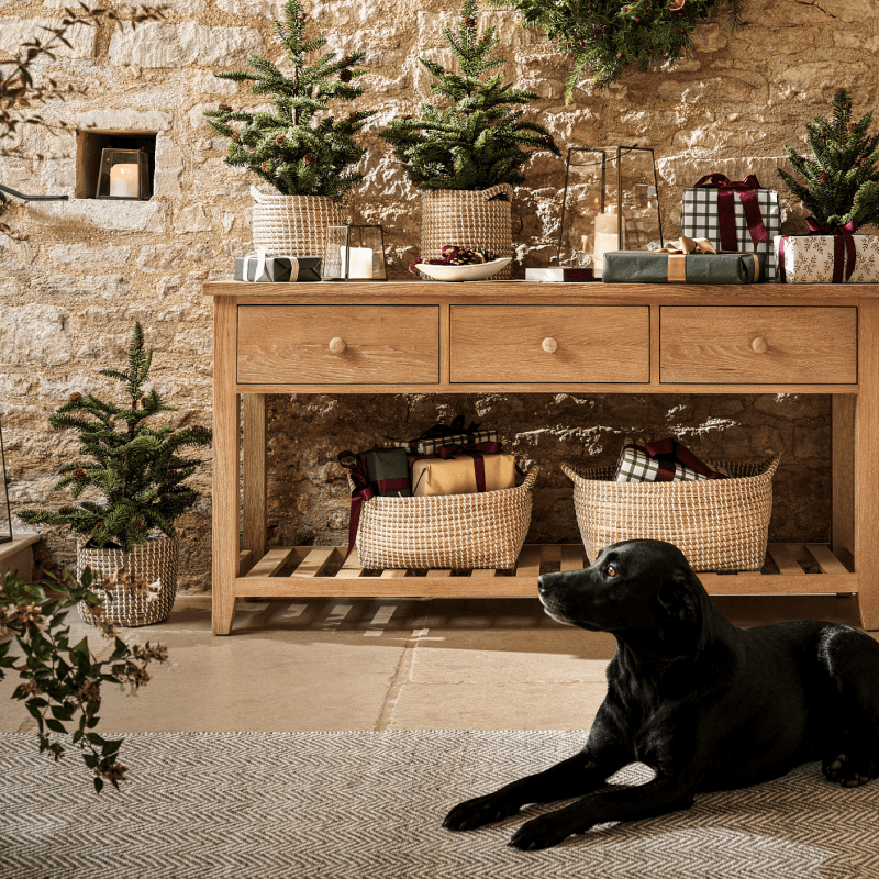 A room with a wooden console table decorated with small pine trees and gifts. A black dog lies on the floor, and a stone wall with a candle niche.