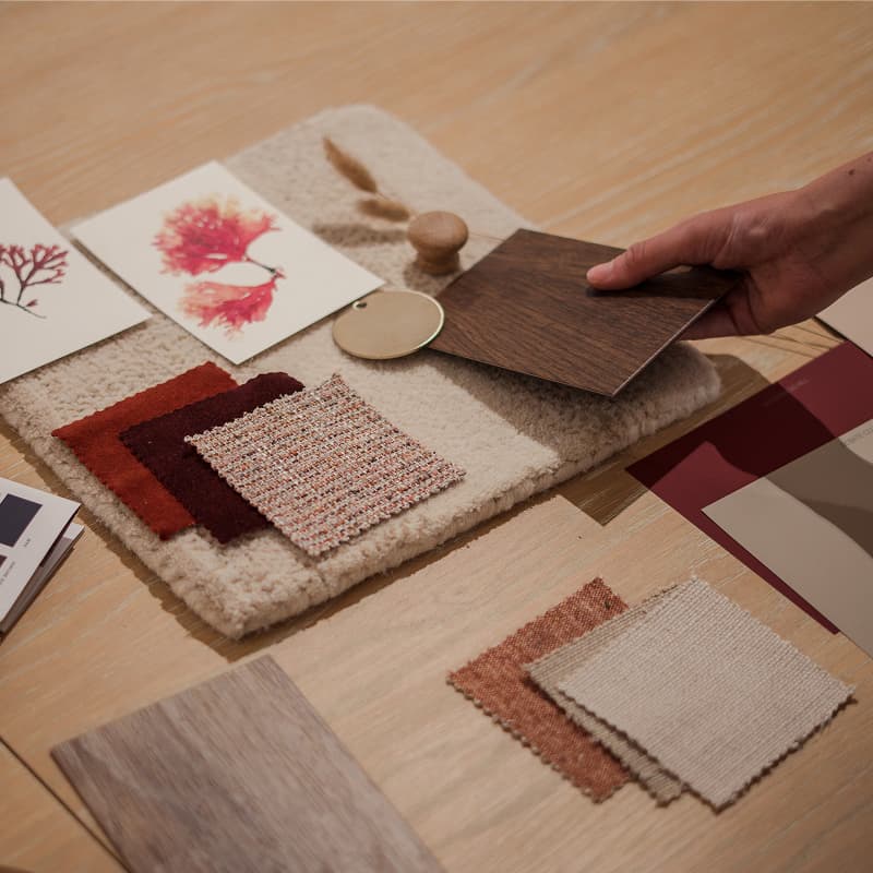 A hand holds a wood sample over a table with fabric swatches, paint chips, dried flowers, and design cards.