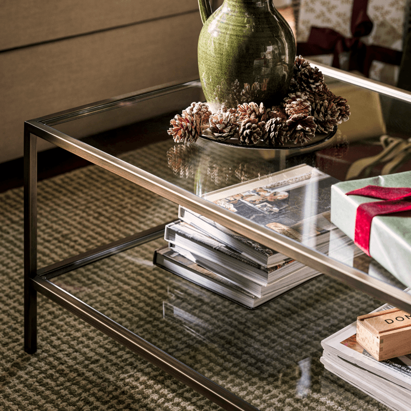 A glass coffee table with a green vase and pine cones, magazines, and a wrapped gift with a red ribbon on a checkered carpet.