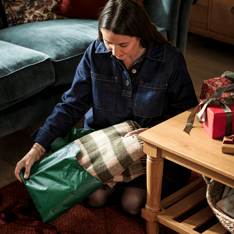 A woman in a denim outfit unwrapping a green gift, revealing a plaid blanket, sitting next to a table with wrapped presents.