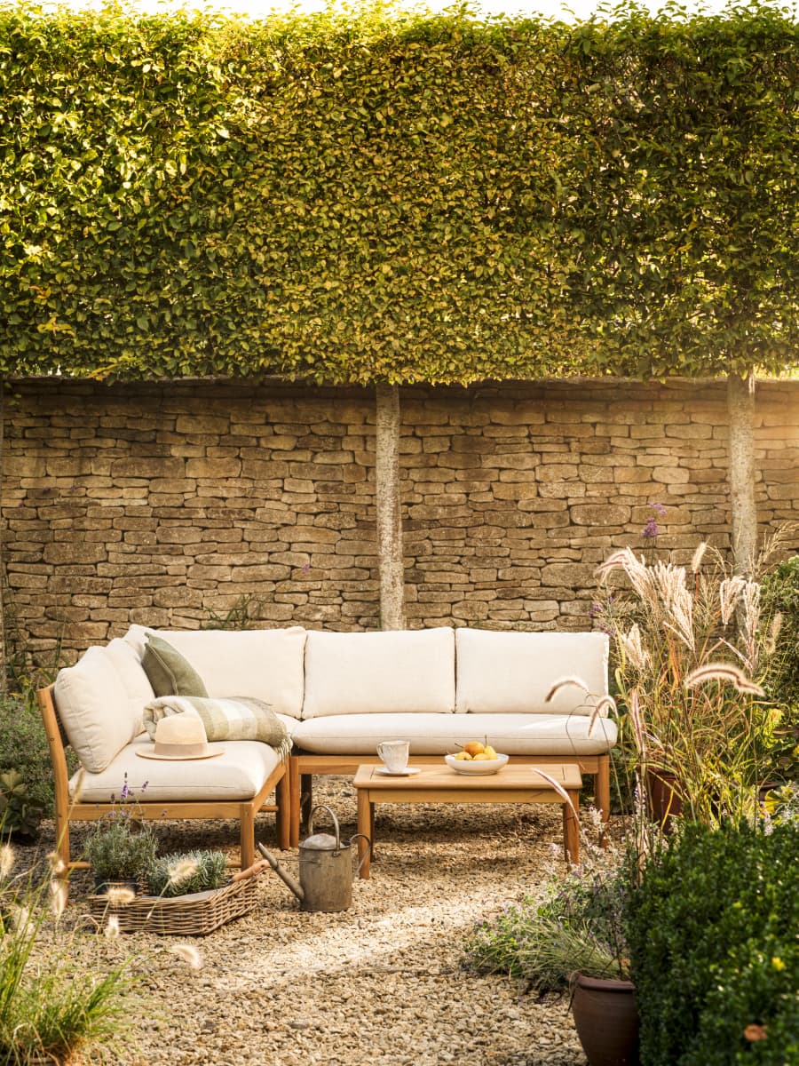 Outdoor patio with cream sectional sofa, coffee table, basket with plants, watering can, and a hat. Stone wall and greenery in the background.