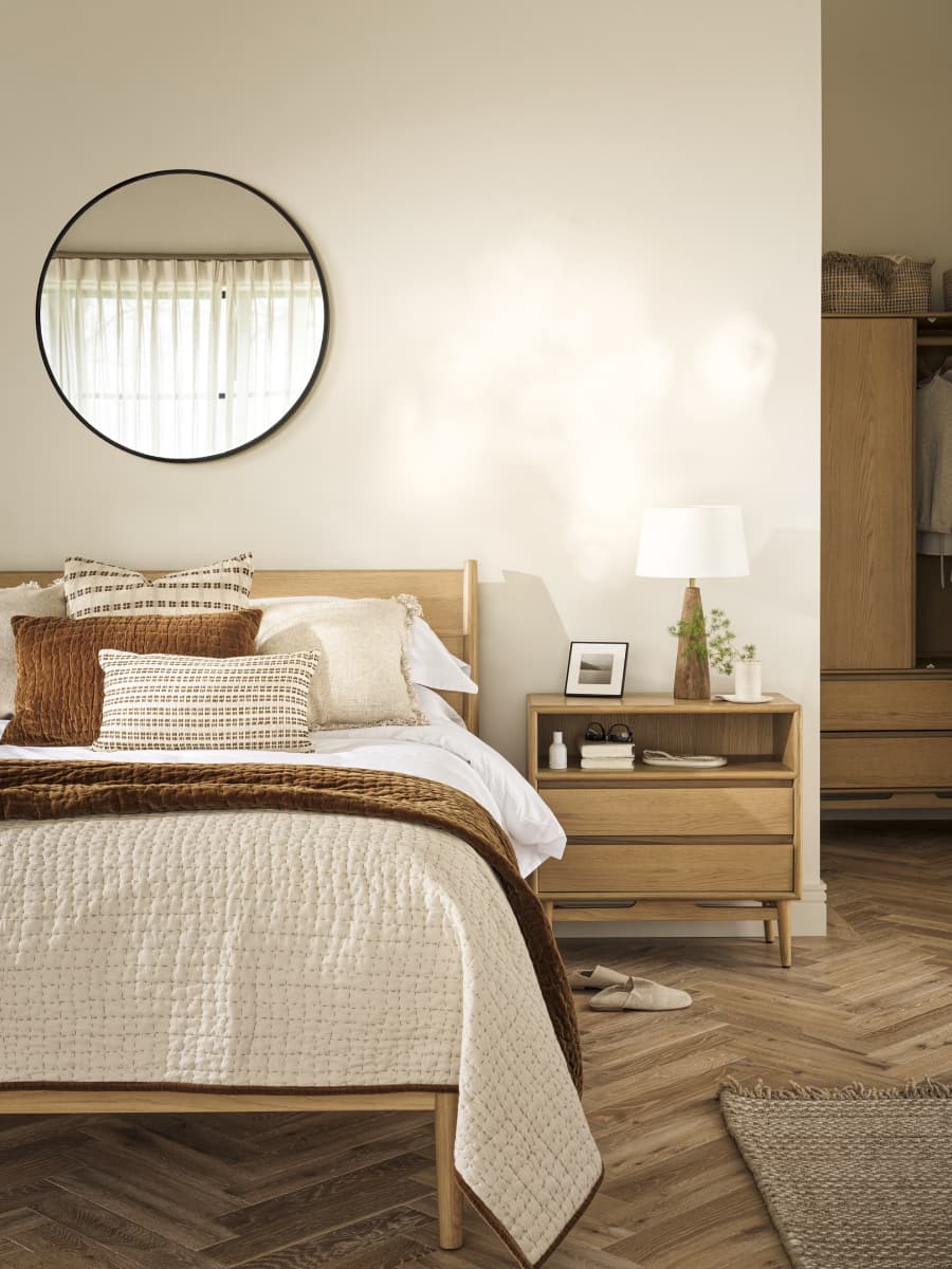 Cozy bedroom with a wooden bed, layered cushions, and a quilt. A round mirror hangs above, next to a nightstand with a lamp and decor.