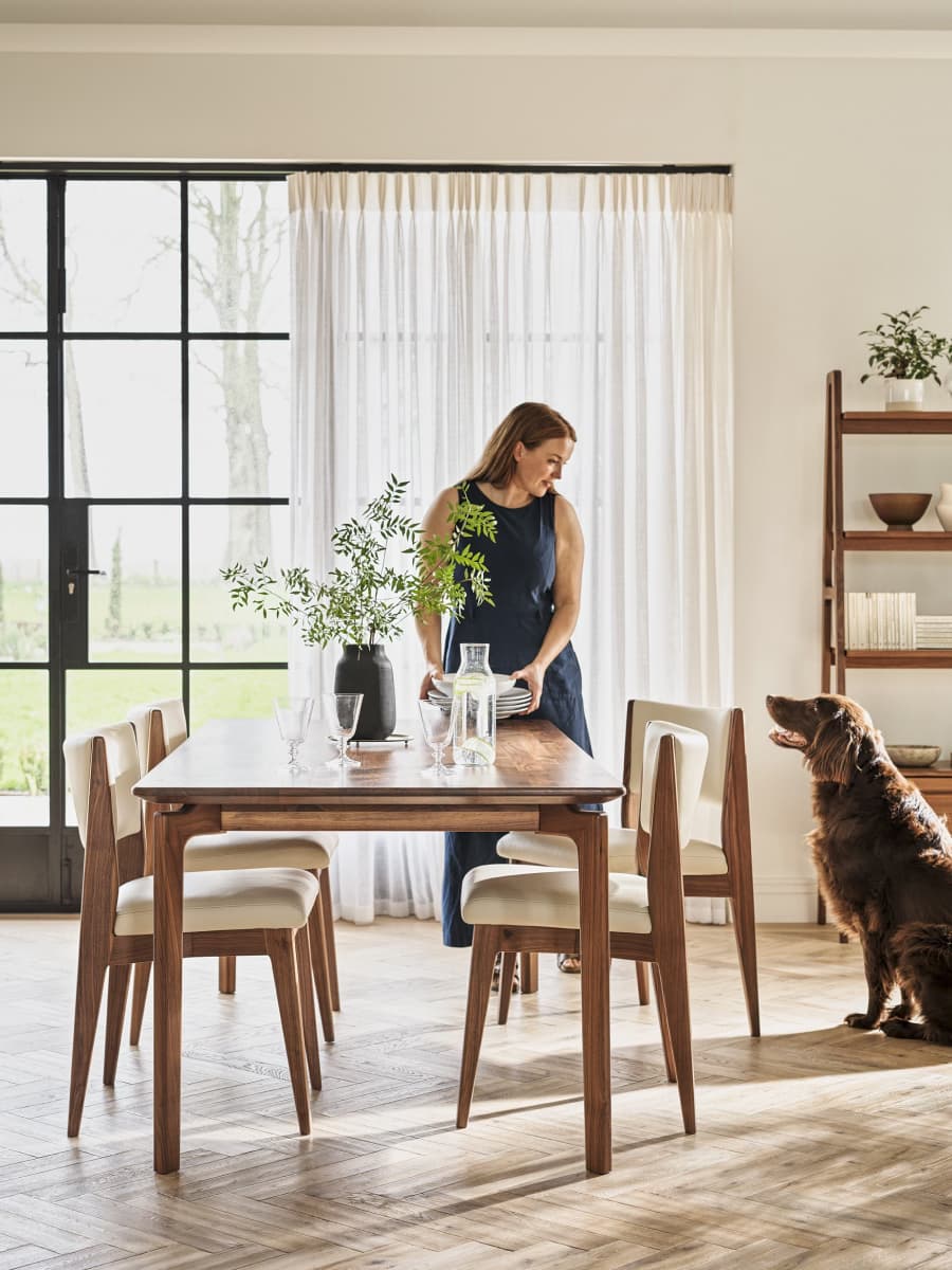 A woman sets a wooden dining table with plates and glasses in a sunlit room, as a brown dog looks on.