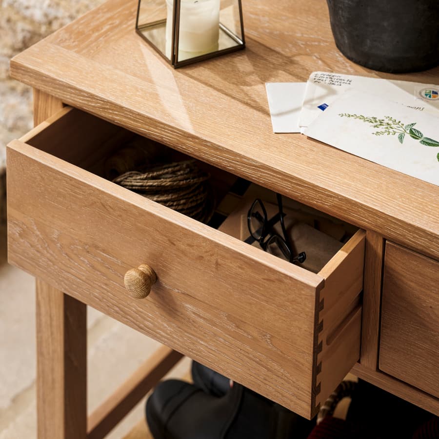 A wooden table with an open drawer containing a pair of glasses and rope, with letters, a candle, and a potted plant on top.