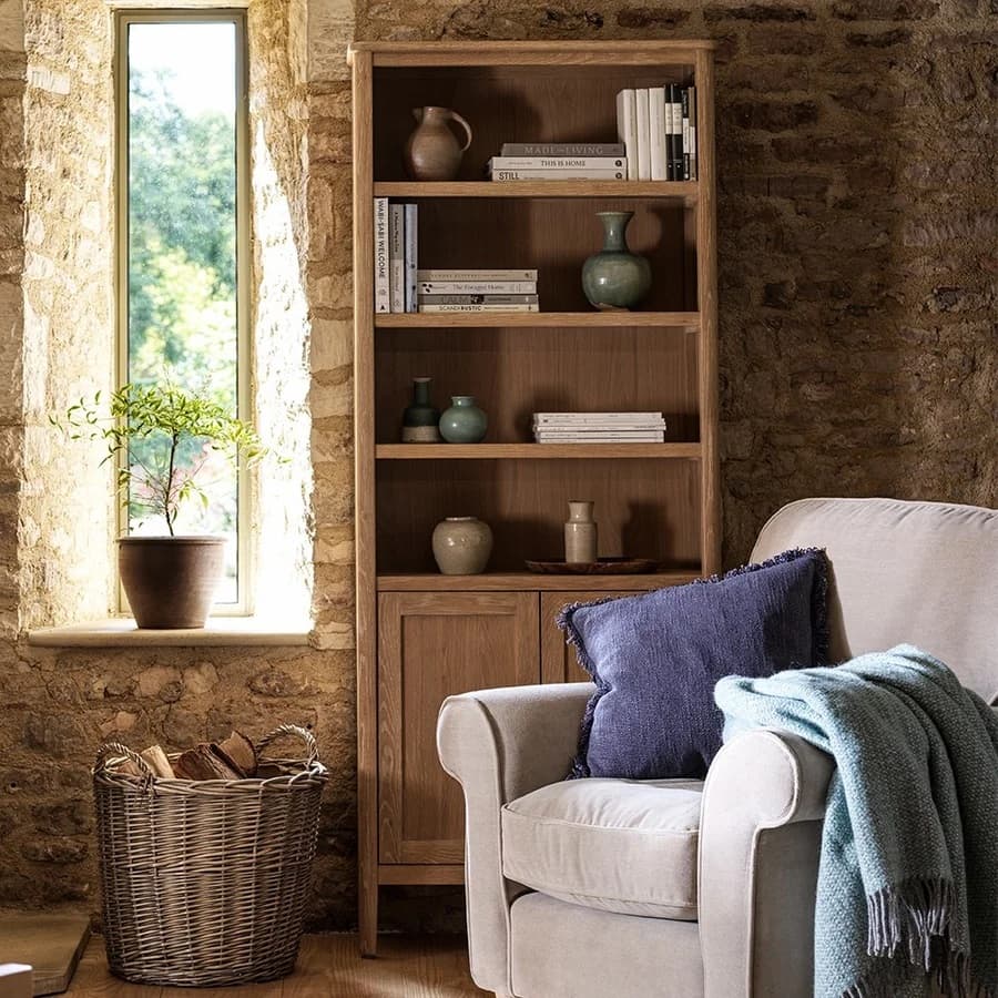 Room with a stone wall, tall bookshelf, cream armchair, blue cushion, teal throw, potted plant by window, and wicker basket with logs.