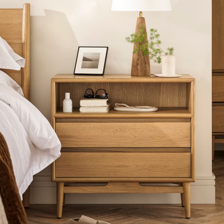 Wooden bedside table with two drawers, a lamp, framed photo, books, and glasses. Nearby a bed with white linens.