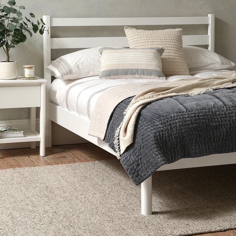 A bedroom with a white bed, layered pillows and blankets in neutral tones, a wooden nightstand with a potted plant, and a beige area rug.